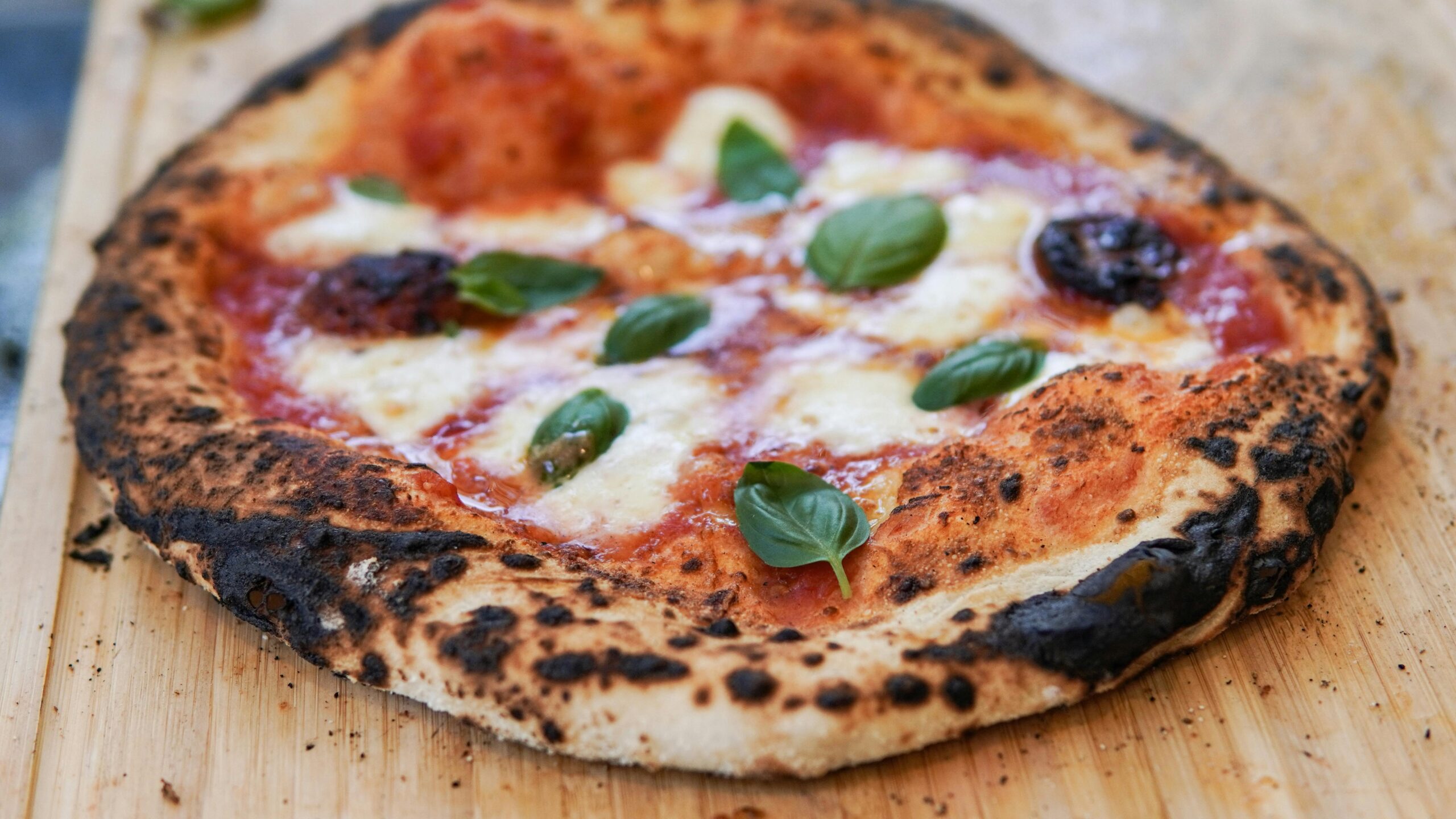 Close-up of a wood-fired Margherita pizza with fresh basil on a wooden surface.